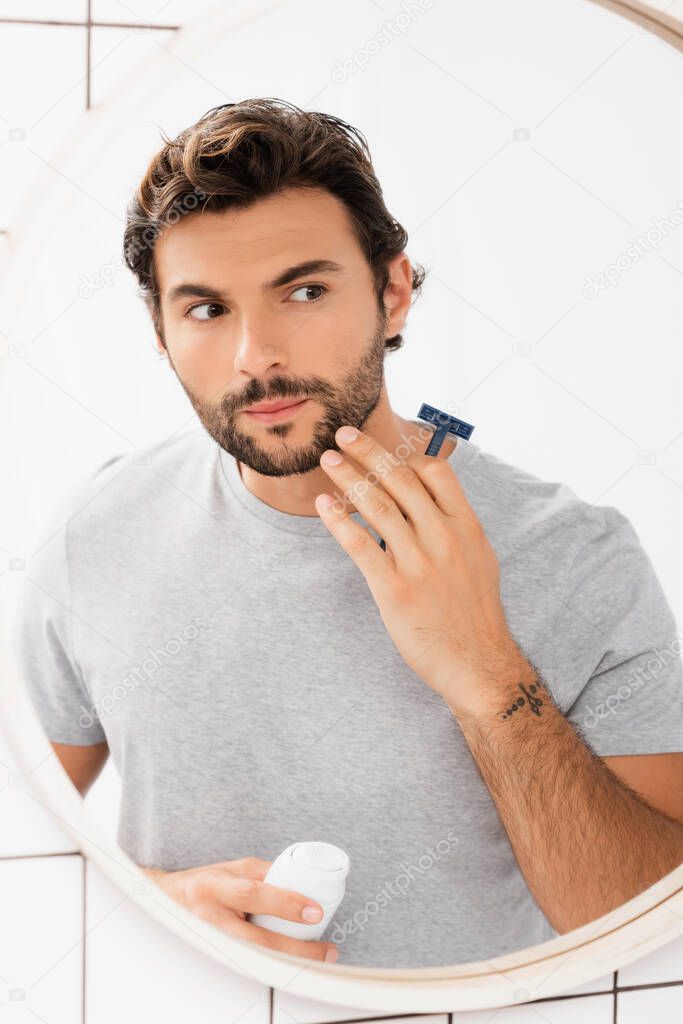 Bearded man touching chin while holding razor and shaving foam near mirror in bathroom 