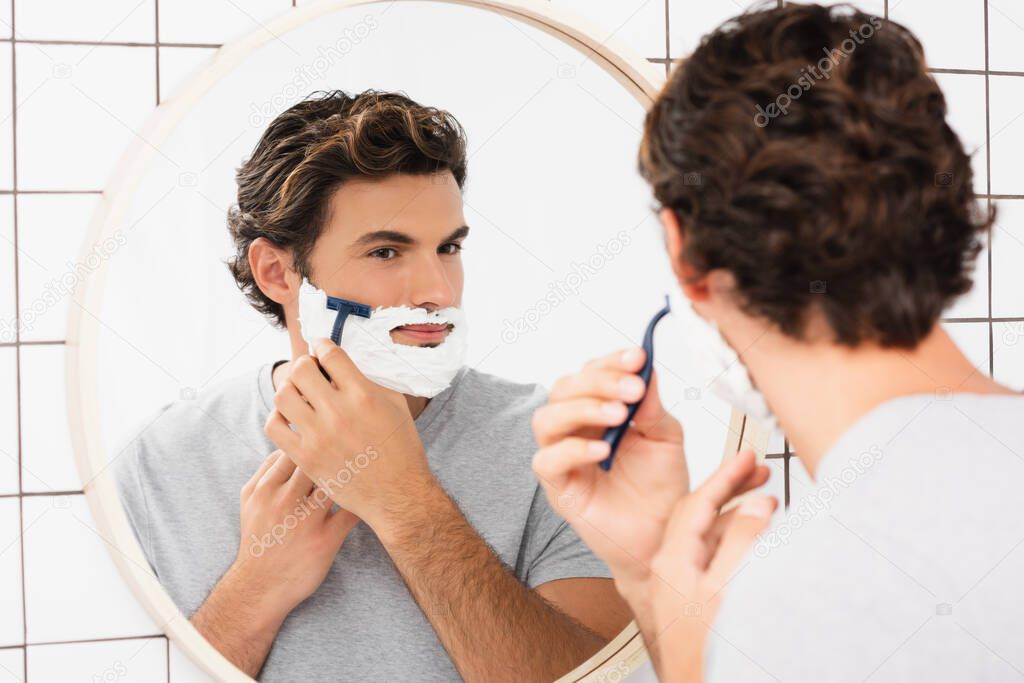 Young man looking at mirror while shaving on blurred foreground in bathroom 