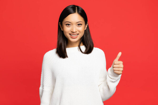 happy asian woman in sweater showing thumb up isolated on red 