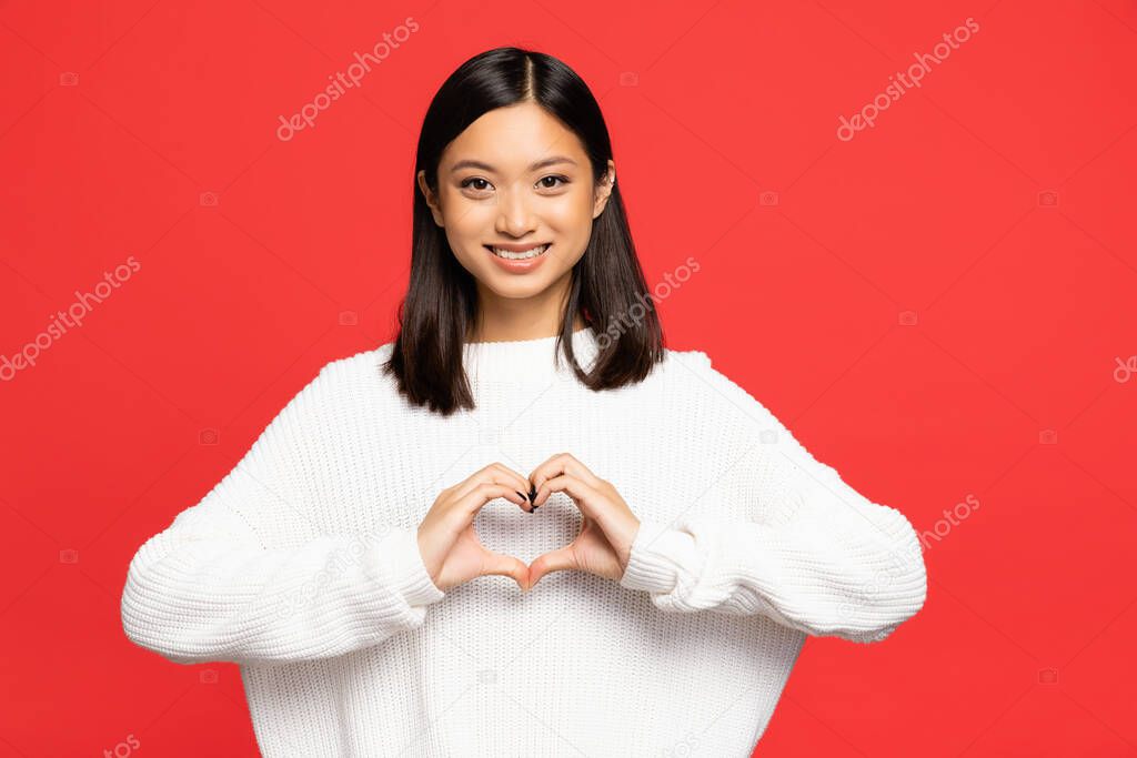 happy and young asian woman showing heart sign with hands isolated on red