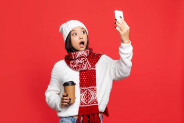Kırmızı renkte selfie çekerken elinde kahve tutan şapkalı ve fularlı Asyalı kadın şok oldu.