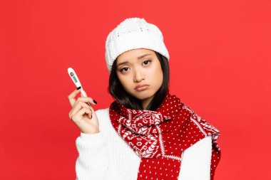 sad asian woman in hat and scarf holding electronic thermometer isolated on red  clipart