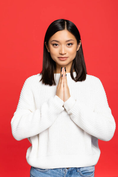 happy and young asian woman standing with praying hands isolated on red