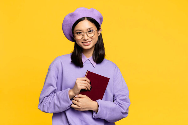 happy asian woman in beret and eyeglasses holding notebook isolated on yellow 
