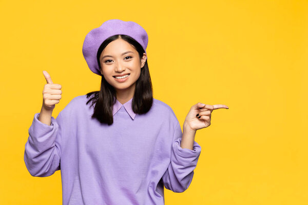 счастливая азиатская женщина в берете показывает большой палец вверх и указывая пальцем изолированы на желтый