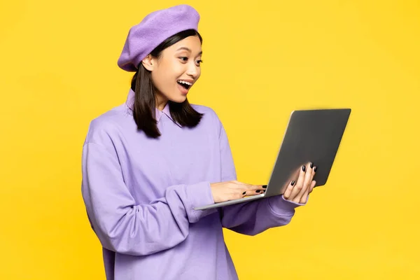 快乐的亚洲女人在贝雷帽使用笔记本电脑隔离黄色 — 图库照片