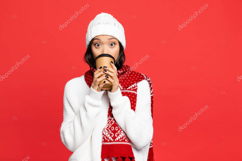surprised asian woman in hat and scarf drinking coffee to go isolated on red