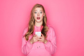 Schockierte schöne Frau mit offenem Mund hält Smartphone auf rosa Hintergrund