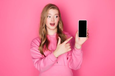Güzel bir kadın pembe arka planda akıllı telefonu işaret ediyor.