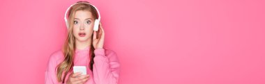 Şok olmuş, kulaklıklı, pembe arka planda akıllı telefonlu güzel bir kadın.