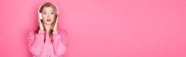 Şok olmuş güzel bir kadın pembe arka planda kulaklıkla müzik dinliyor.
