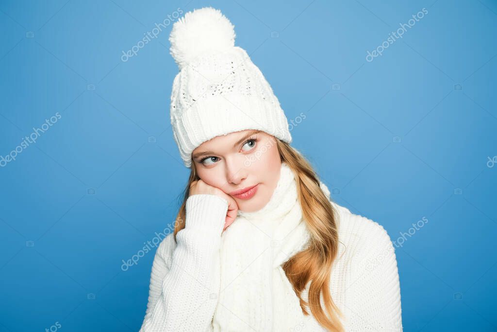 pensive blonde beautiful woman in winter white outfit on blue background