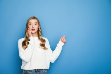 shocked blonde beautiful woman in sweater pointing with finger isolated on blue clipart
