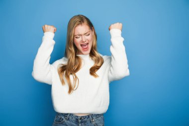 happy blonde beautiful woman in sweater showing yeah gesture isolated on blue background clipart