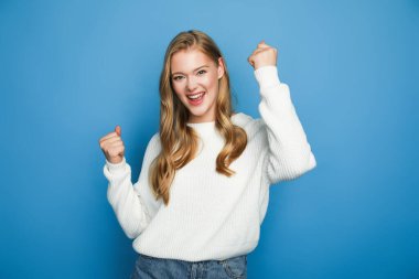 happy blonde beautiful woman in sweater showing yeah gesture isolated on blue background clipart