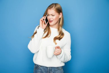 confused blonde woman in sweater talking on smartphone with mug isolated on blue background clipart