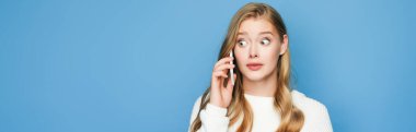 Mavi arka planda izole edilmiş akıllı telefondan konuşan süveterli sarışın güzel bir kadın.