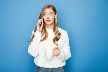 Süveter giyen sarışın güzel bir kadının mavi arka planda izole edilmiş bir kupayla akıllı telefondan konuşmasına şaşırdım.