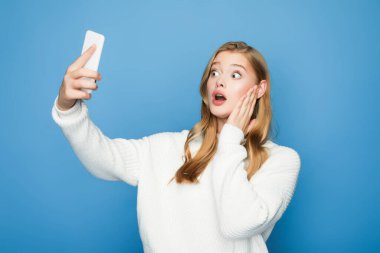 Şok olmuş sarışın güzel kadın mavi arka planda selfie çekiyor.