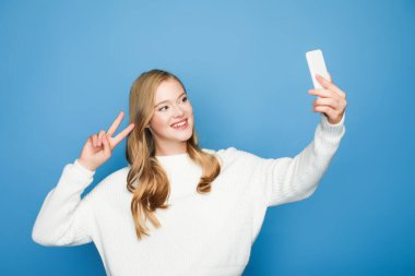 Sarışın güzel kadın selfie çekiyor ve mavi arka planda izole edilmiş barış işareti gösteriyor.