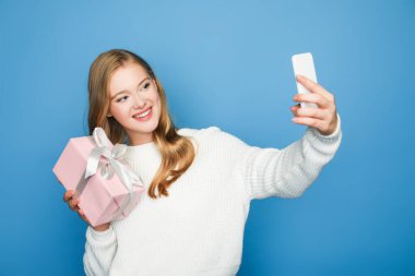 Süveter giymiş sarışın güzel bir kadın mavi renkli bir hediye kutusuyla selfie çekiyor.