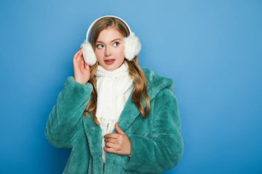 stylish woman in green faux fur coat and ear muffs isolated on blue clipart