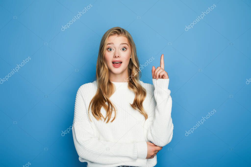 excited blonde beautiful woman in sweater showing idea gesture isolated on blue