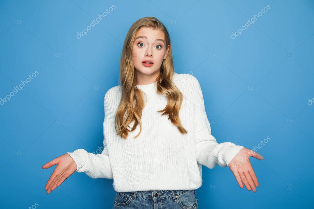 surprised blonde beautiful woman in sweater showing shrug isolated on blue background
