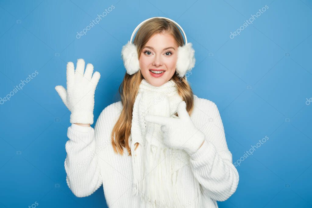 smiling beautiful woman in winter white outfit pointing at hand in glove isolated on blue