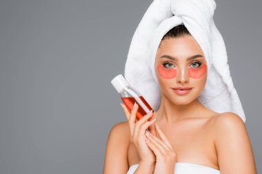 woman with towel on head and eye patches holding lotion isolated on grey clipart