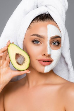 Kafasında havlu ve yüzünde kilden maske olan kadın gri üzerinde avakado tutuyor.