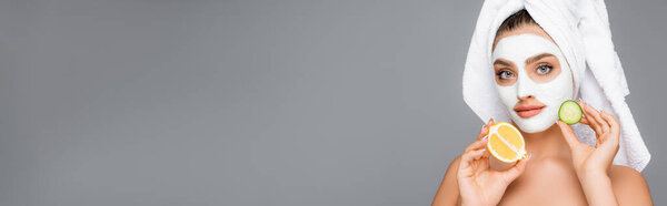 woman with towel on head and clay mask on face holding lemon and cucumber isolated on grey, banner