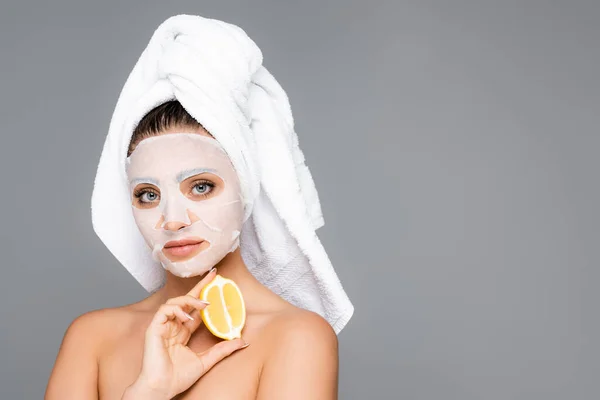 woman with towel on head and mask sheet on face holding lemon isolated on grey