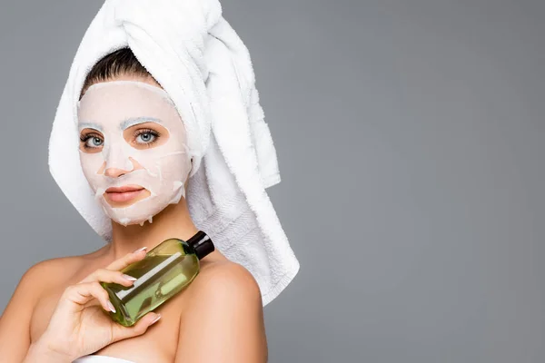 woman with towel on head and mask sheet on face holding bottle isolated on grey