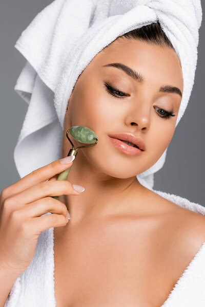 woman with towel on head using jade roller on face isolated on grey