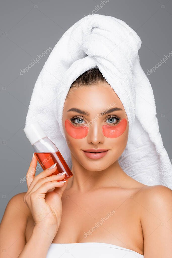 woman with towel on head and eye patches holding lotion isolated on grey