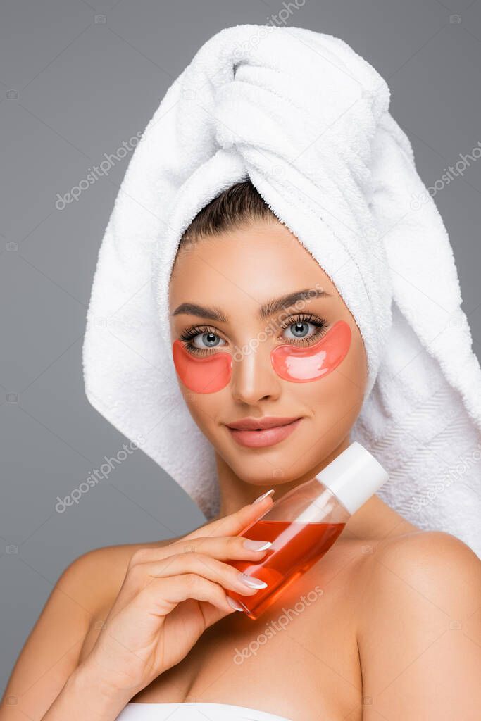 woman with towel on head and eye patches holding lotion isolated on grey