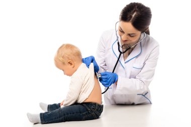 Gülümseyen bir çocuk doktoru, steteskopu beyaza ayrılmış bir çocuğun arkasını inceliyor.