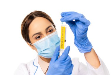 Tıbbi maskeli doktorun seçici odak noktası elinde kovid 19 aşısı olan bir şırınga. Beyaz harflerle izole edilmiş.
