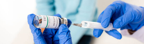 Panoramic crop of doctor in latex gloves holding syringe and vaccine with coronavirus lettering isolated on white