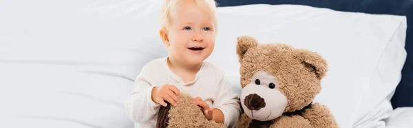 パノラマショットの笑顔幼児男の子座っています近くに柔らかいおもちゃオンベッド — ストック写真