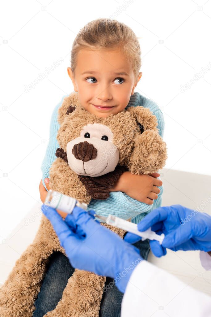 Selective focus of smiling girl holding soft toy near pediatrician with vaccine and syringe isolated on white