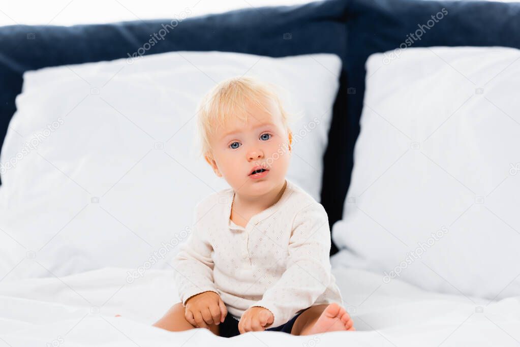 Selective focus of toddler boy looking at camera on bed on white background 