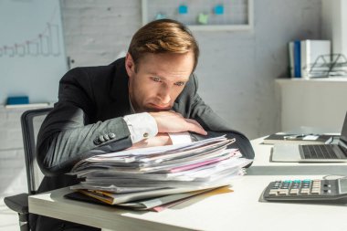 Disappointed businessman leaning on pile of papers, while sitting at workplace on blurred background clipart