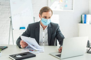 Tıbbi maskeli bir iş adamı, elinde kağıtlar ve dizüstü bilgisayarla, arka planda bulanık bir şekilde işyerinde oturuyor.