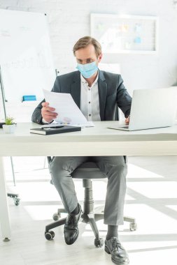 Tıbbi maske takmış bir iş adamı masasında otururken kağıda bakar.