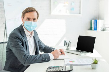 Businessman in medical mask looking at camera, while writing in notebook at workplace on blurred background clipart
