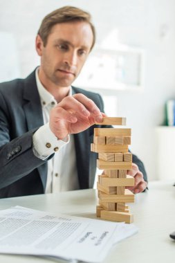 Focused businessman building blocks wood game, while sitting at workplace with blurred petition for bankruptcy on foreground clipart