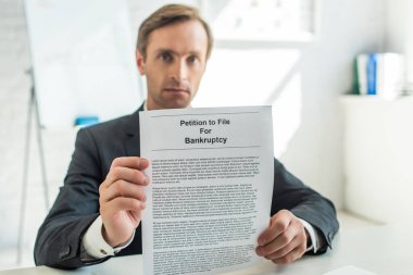 Serious businessman looking at camera and showing petition for bankruptcy, while sitting at desk on blurred background clipart