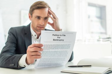 Pensive businessman looking at petition for bankruptcy, while sitting at workplace on blurred background clipart
